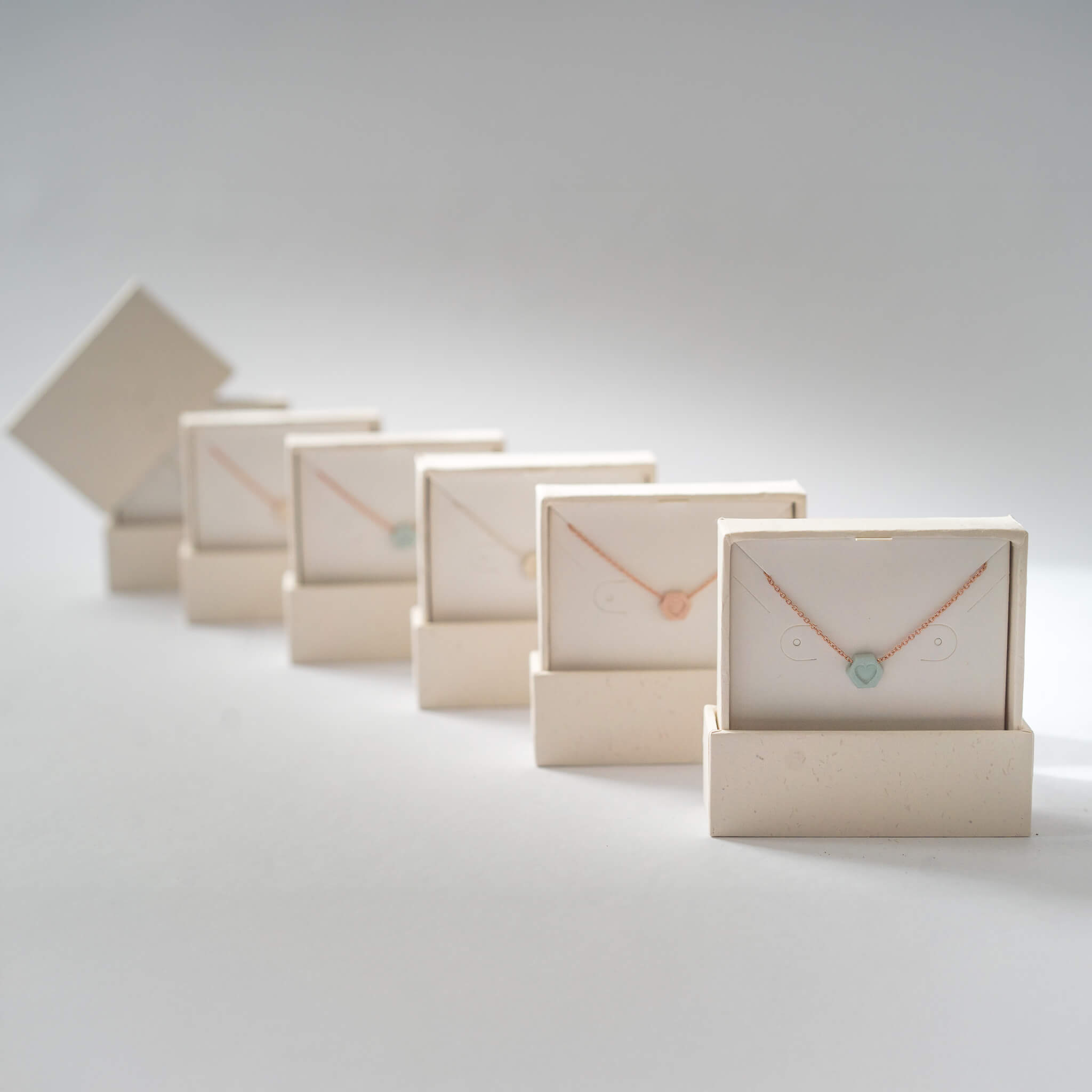 Collection of handcrafted porcelain initial necklaces in ivory, pink, and blue displayed in eco gift boxes.